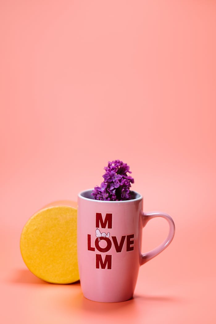 Pink mug with 'Love Mom' text filled with purple flowers on pink background.