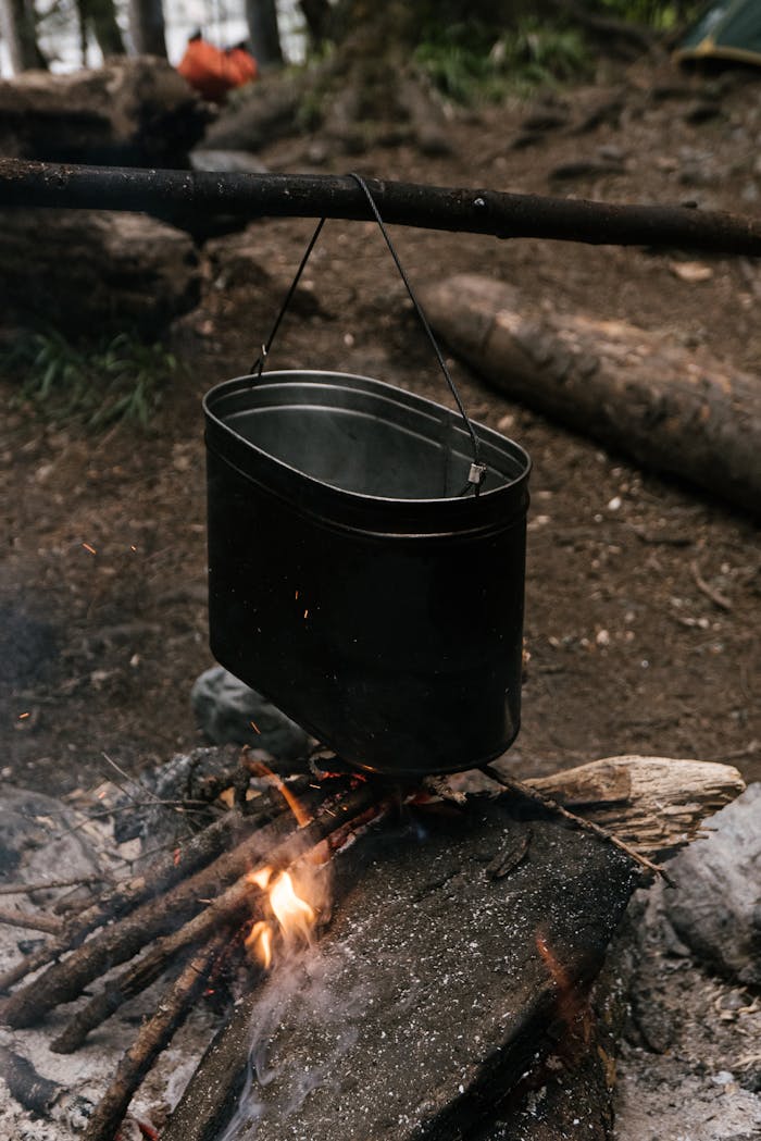 A tin container hangs over a campfire in a rustic outdoor camping setup.